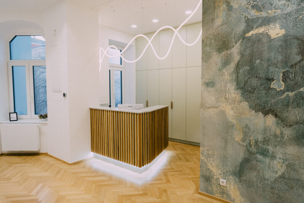 Empfangstresen mit Holzlatten in modernem Innenraum, umgeben von großen Fenstern und gemalter Wand mit abstraktem Muster. Hölzerner Boden ergänzt die helle, minimalistische Einrichtung.