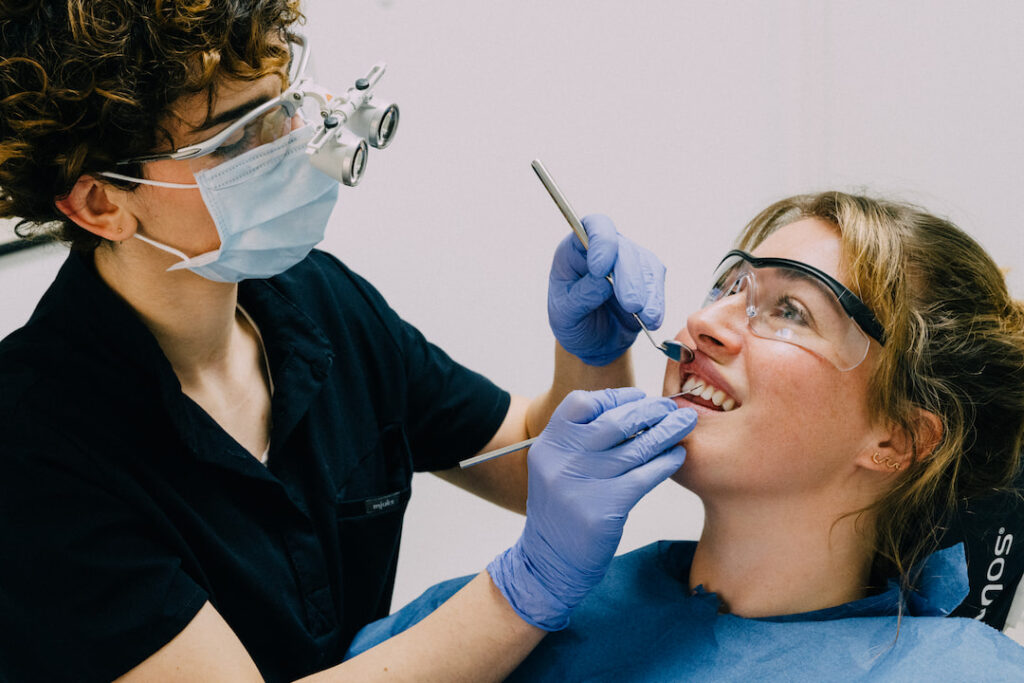 Zahnärztin untersucht Patientin mit Zahnarztspiegel und Schutzbrille in Praxis. Beide tragen Schutzbrillen, Arzt trägt OP-Maske und Handschuhe.